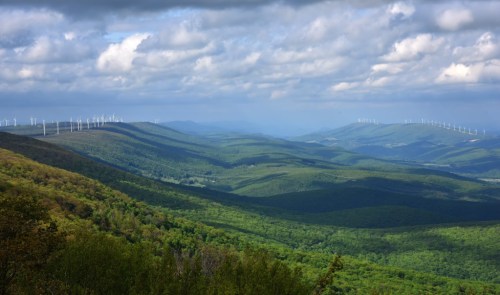 A panoramic view of rolling green hills under a cloudy sky, with wind turbines visible in the distance.