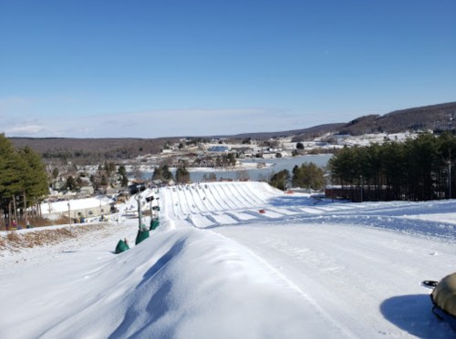 A snowy landscape with a tubing hill, trees, and a view of a lake and town in the distance under a clear blue sky.