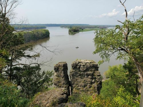 A scenic view of a river winding through lush greenery, with rocky cliffs in the foreground and a small boat on the water.