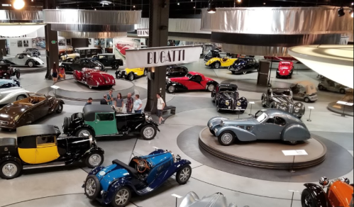 A spacious car museum featuring various vintage cars, including Bugatti models, with visitors exploring the exhibits.