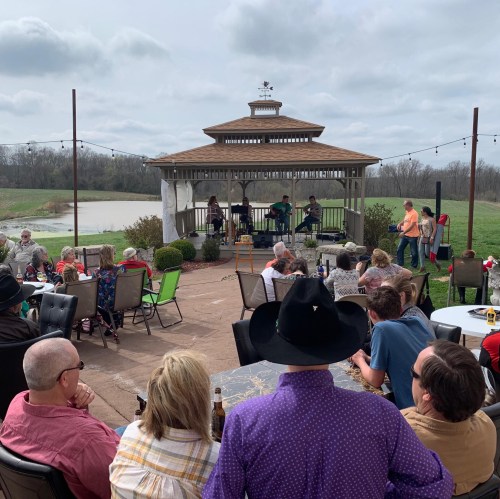 A crowd enjoys live music at a gazebo by a lake, with people seated at tables and a cloudy sky overhead.