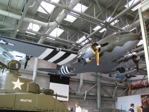 A vintage military aircraft displayed in a museum, with a tank in the foreground and flags in the background.