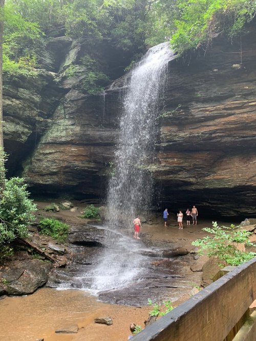 A waterfall cascades down rocky cliffs, with people standing in the shallow water below, surrounded by lush greenery.