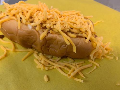 A hot dog topped with a generous amount of shredded cheddar cheese on yellow paper.