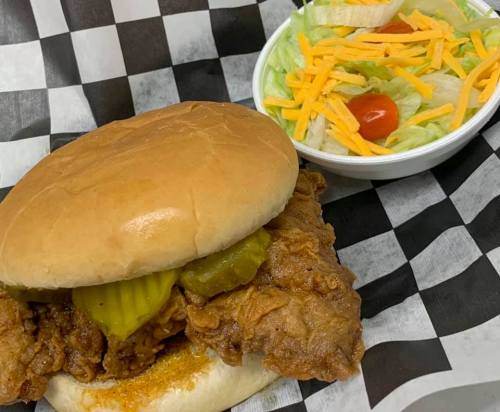 A crispy fried chicken sandwich with pickles on a bun, served with a side salad of lettuce, cheese, and tomato.