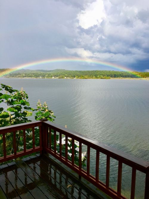 A vibrant rainbow arcs over a calm lake, framed by dark storm clouds and lush greenery.