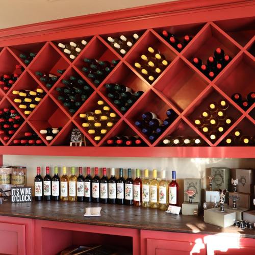 A red wine rack filled with various bottles, alongside a wooden counter displaying more wine bottles and a sign.