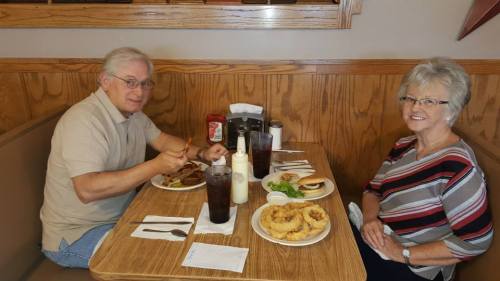 A man and woman enjoy a meal at a restaurant, with burgers, onion rings, and drinks on the table.
