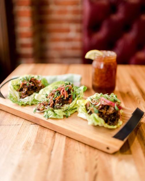 Three lettuce wraps filled with savory meat and topped with herbs, served on a wooden platter with a drink in the background.