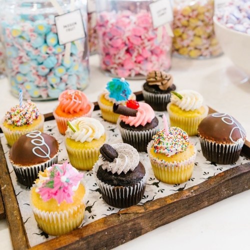 A wooden tray holds a variety of colorful cupcakes, surrounded by jars of candy in the background.
