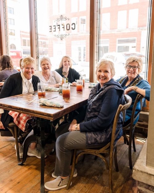 A group of five women smiling and sitting around a wooden table in a cozy café.