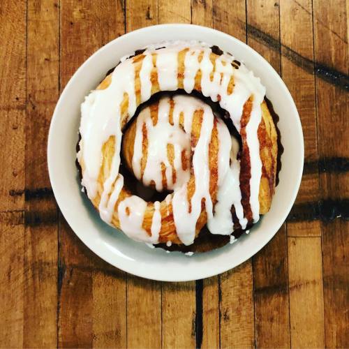 A freshly baked cinnamon roll drizzled with white icing, served in a white bowl on a wooden table.