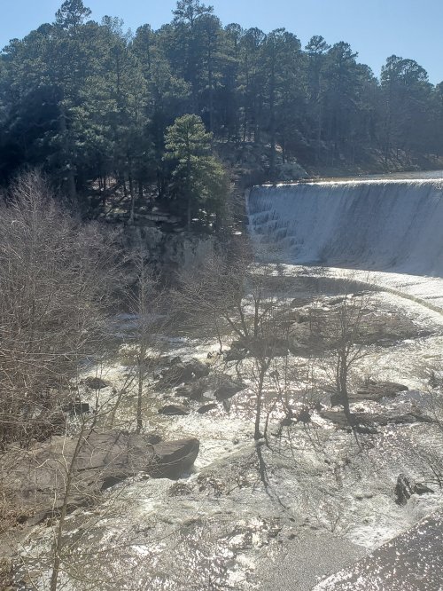 A waterfall cascades over a rocky ledge, surrounded by trees and a clear blue sky.