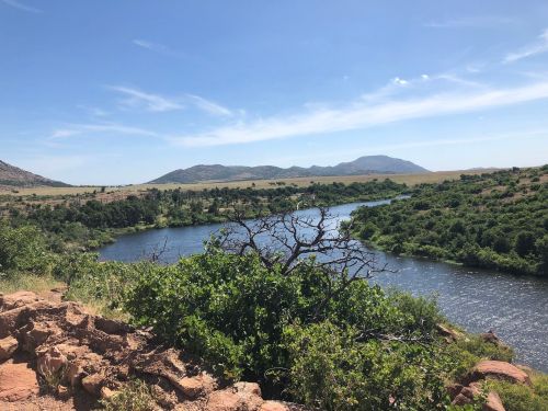 A serene river flows through a lush landscape, surrounded by rolling hills and a clear blue sky.