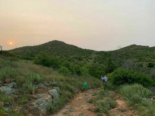 Two people walk along a rocky trail in a green landscape at sunset, with hills and a hazy sky in the background.