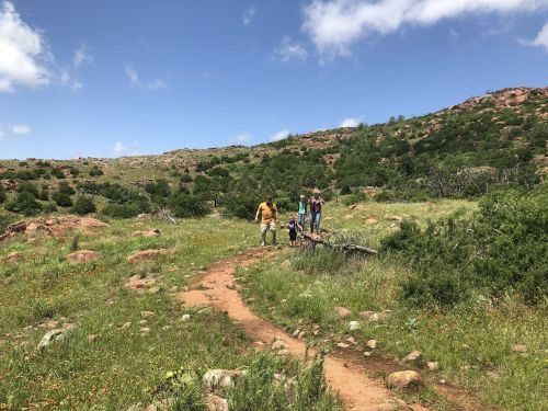 A family walks along a dirt path through a green, hilly landscape with scattered rocks and blue skies.