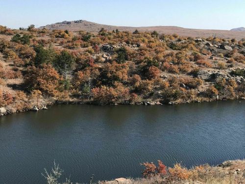 A serene river flows through a landscape of autumn-colored trees and rocky hills under a clear blue sky.