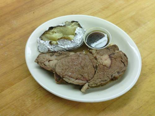 A plate with a slice of roast beef, a baked potato wrapped in foil, and a small cup of sauce.