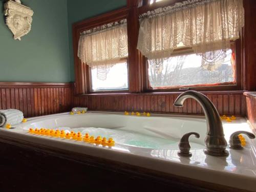 A cozy bathroom with a jetted tub filled with bubbles and lined with rubber duckies, set against wooden paneling and lace curtains.