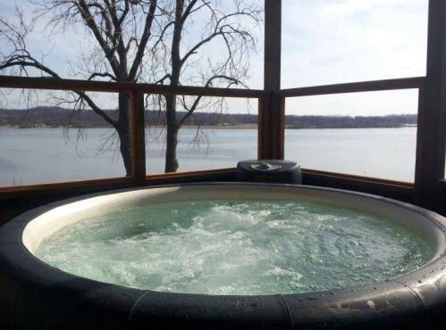 A hot tub with bubbling water, overlooking a serene lake and trees in the background under a clear sky.