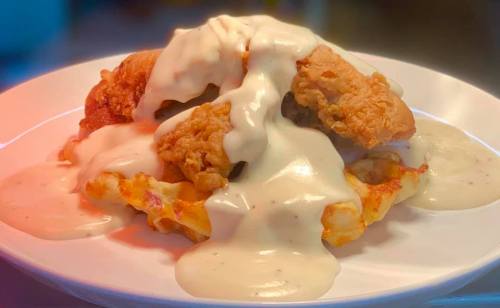 A plate of crispy fried chicken topped with creamy white gravy over a bed of cheesy pasta.