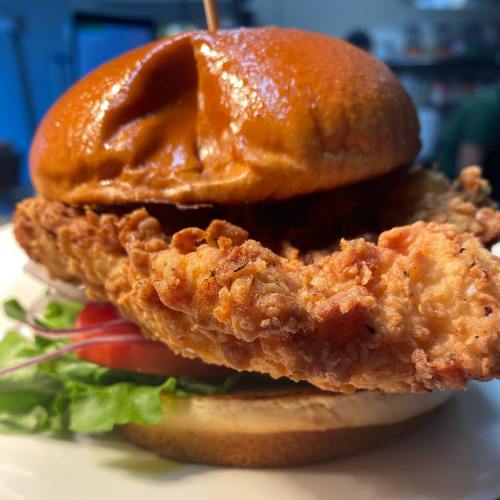 Close-up of a crispy fried chicken sandwich with lettuce, tomato, and a shiny bun on a white plate.