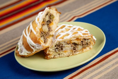 Two savory pastries filled with meat and vegetables, drizzled with white icing, on a green plate against a colorful background.