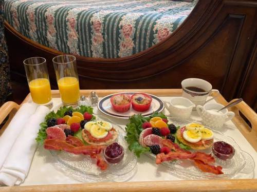A breakfast tray with two plates of eggs, bacon, fruit, muffins, orange juice, and coffee, set on a floral bedspread.