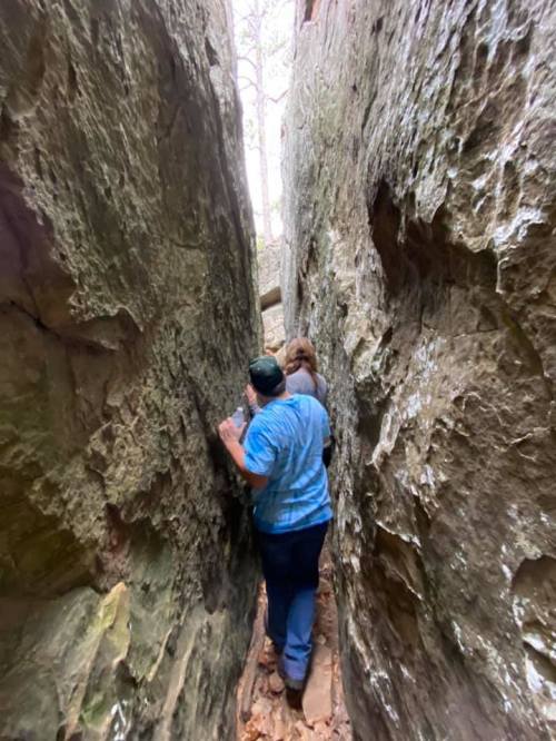 Two people navigate a narrow rock passage in a forested area, surrounded by tall, rugged stone walls.