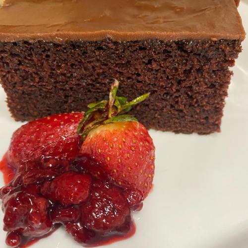 A slice of chocolate cake topped with smooth frosting, accompanied by fresh strawberries and berry sauce.