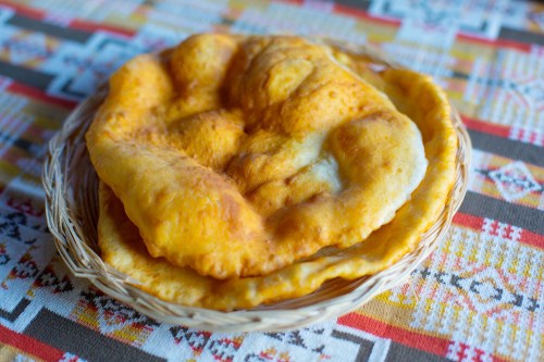 A woven basket holds two golden, fluffy fried breads on a colorful patterned tablecloth.