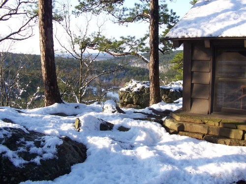 A snowy landscape with a cabin, surrounded by trees and rocky terrain, under a clear blue sky.