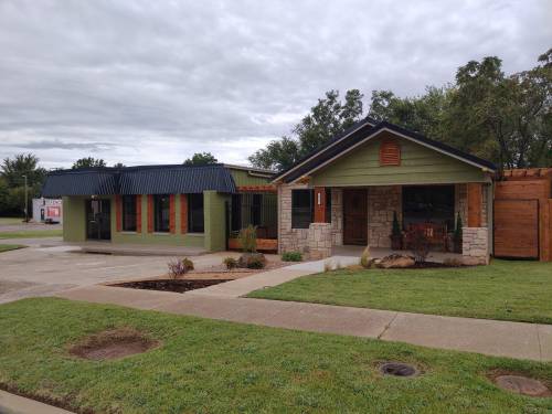 A green and stone building with a wooden porch, next to a modern structure with a metal roof, set in a grassy area.
