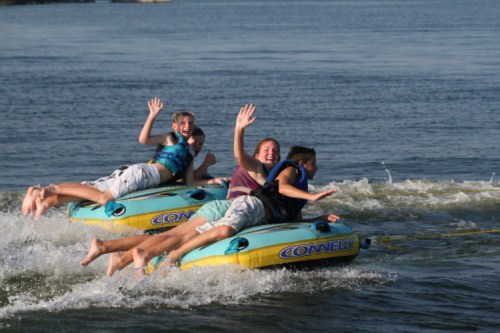 Four people enjoying a fun ride on inflatable tubes being towed on a lake, splashing water and smiling.