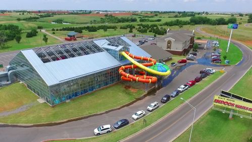 Aerial view of a water park with a large slide, surrounded by green fields and parked cars.