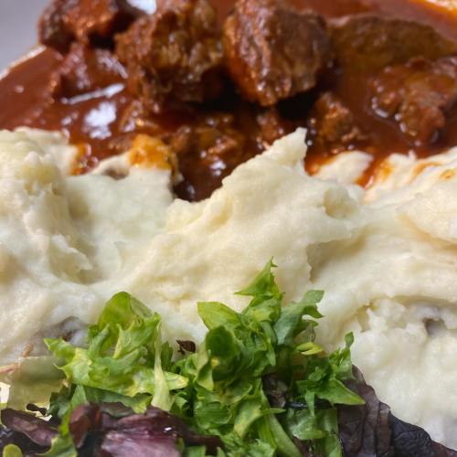 A plate of mashed potatoes topped with green lettuce, accompanied by chunks of meat in a rich red sauce.