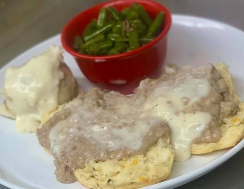 A plate of biscuits topped with creamy sausage gravy, served with a side of green beans in a red bowl.