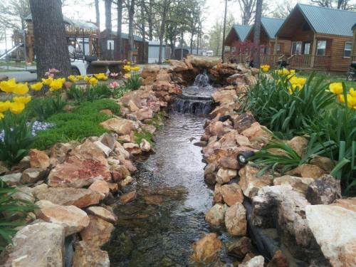 A serene garden scene featuring a small stream surrounded by colorful flowers and rustic cabins in the background.