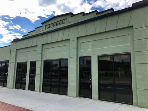Green building with large windows and the word "MONROE" above the entrance, under a blue sky with clouds.