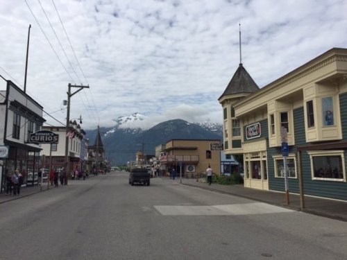A quiet street scene with shops on either side, mountains in the background, and a cloudy sky above.