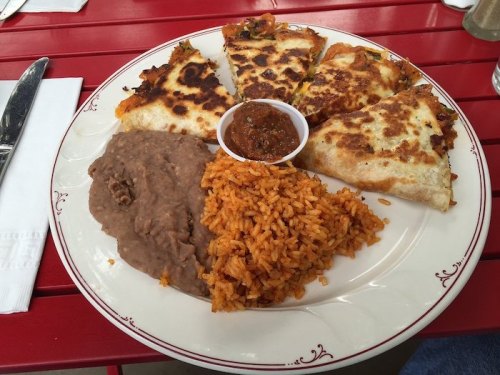 A plate of quesadillas, refried beans, and Spanish rice, served with a small cup of salsa on a red table.
