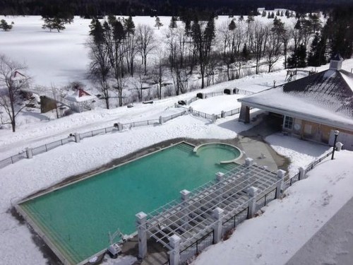A snowy landscape featuring a partially frozen pool and a nearby building, surrounded by trees and a distant lake.