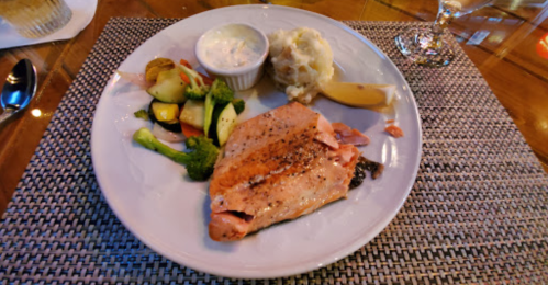 A plate with grilled salmon, mixed vegetables, mashed potatoes, and a small cup of sauce, on a textured placemat.