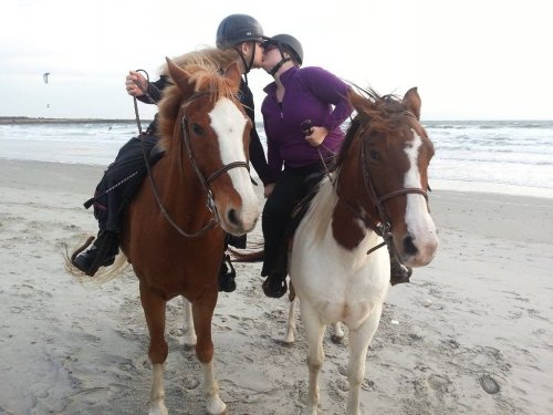 Two women on horseback share a kiss on a beach, with waves and a cloudy sky in the background.