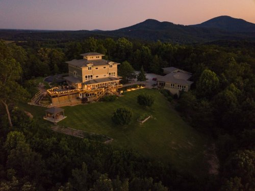 Aerial view of a large house with outdoor lighting, surrounded by trees and mountains at dusk.