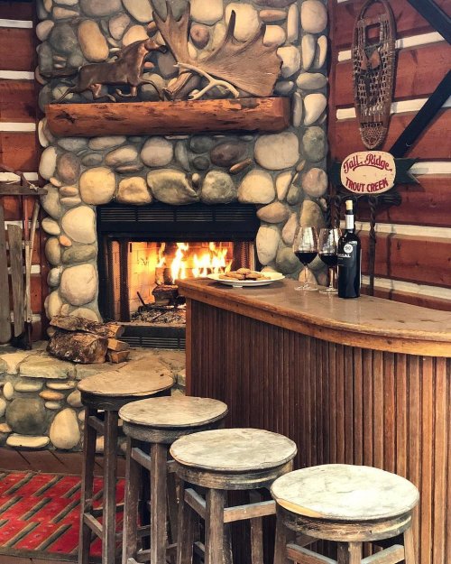 Cozy cabin interior with a stone fireplace, wooden bar, and stools, featuring a bottle of wine and glasses.