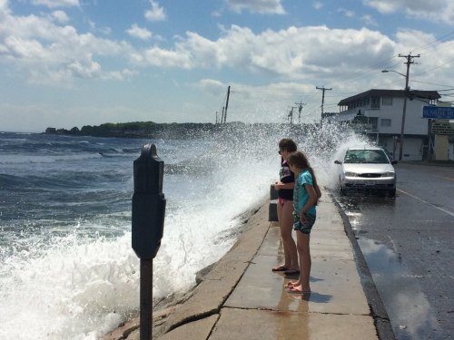 Two girls stand by the shore, watching waves crash against the road on a sunny day with clouds in the sky.