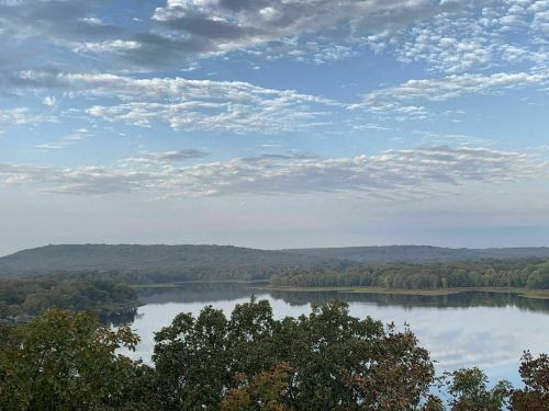 A serene landscape featuring a calm lake surrounded by trees and rolling hills under a partly cloudy sky.
