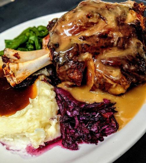 A plate of tender meat with gravy, mashed potatoes, red cabbage, and greens, served on a white dish.