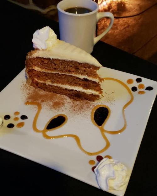 A slice of layered cake with cream frosting on a decorative plate, accompanied by a cup of coffee.